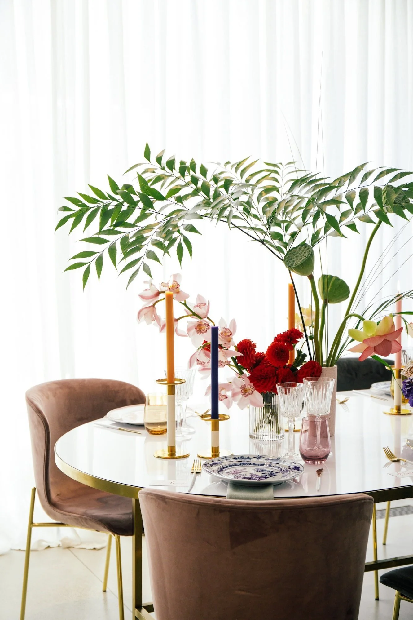 Dining table arranged with greenery, candles and glassware beside sheer curtains.