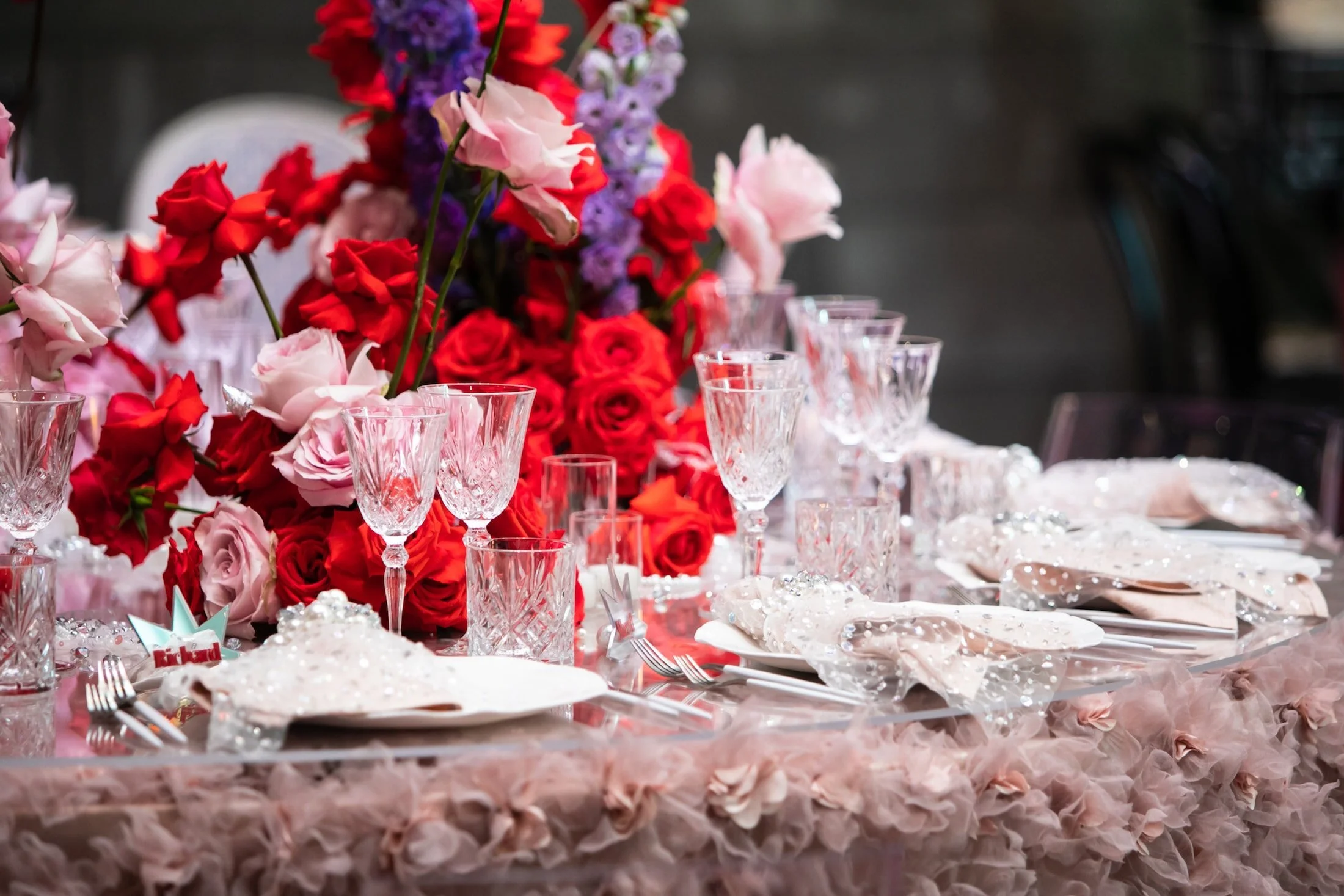 Layered pink table setting with red flowers and clear glassware.