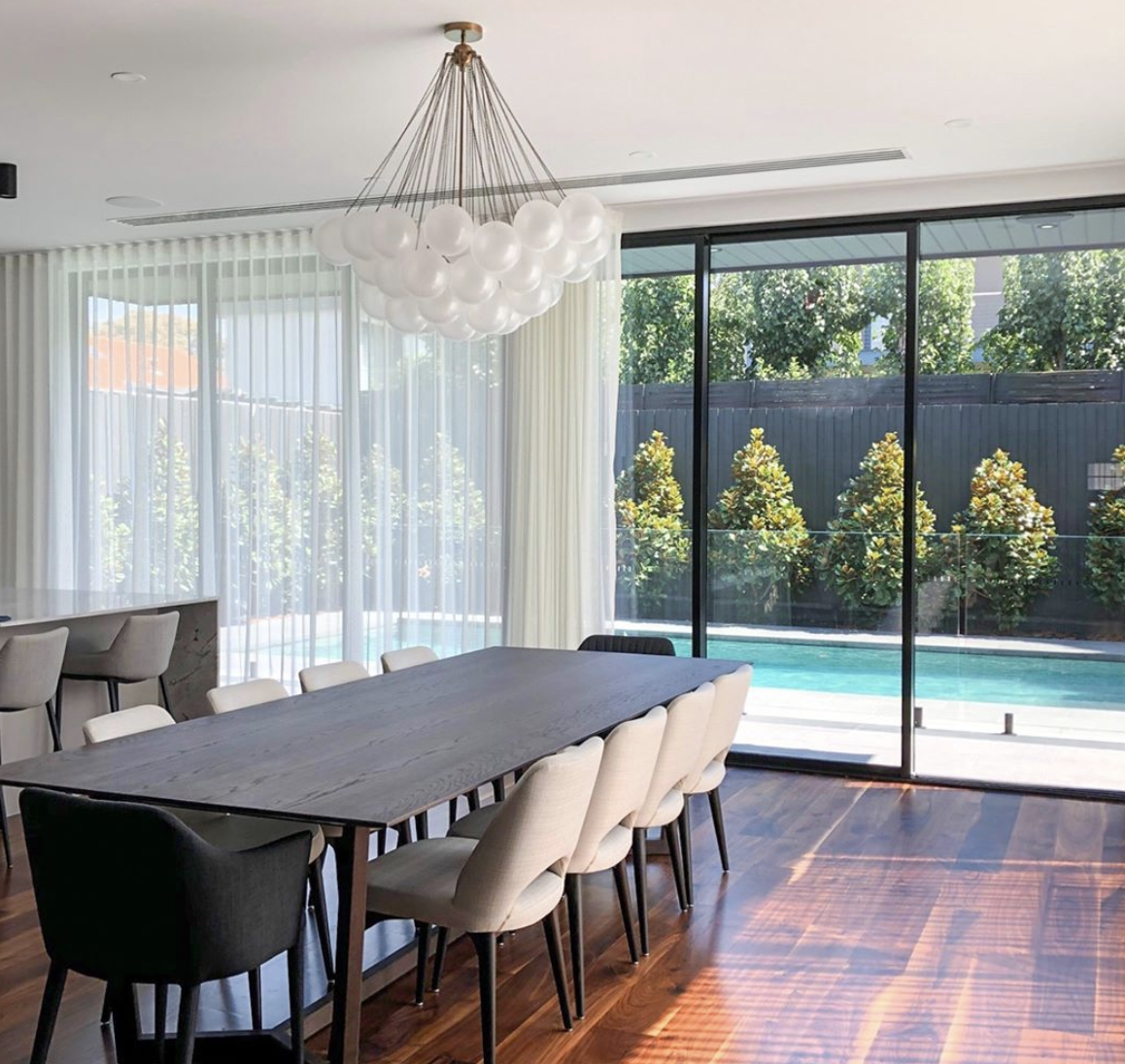 Dining table beneath a decorative pendant beside glazing overlooking a pool.