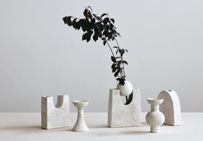 Sculptural pale vessels arranged with a leafy branch against a neutral background.
