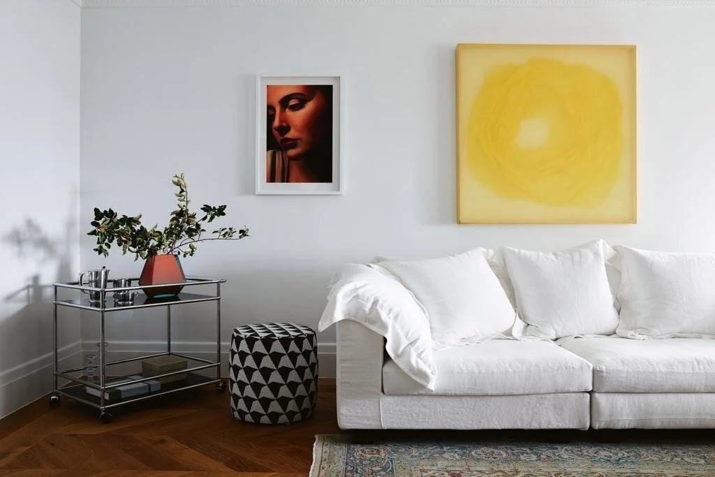 White sofa beneath a yellow artwork beside a glass shelving unit.