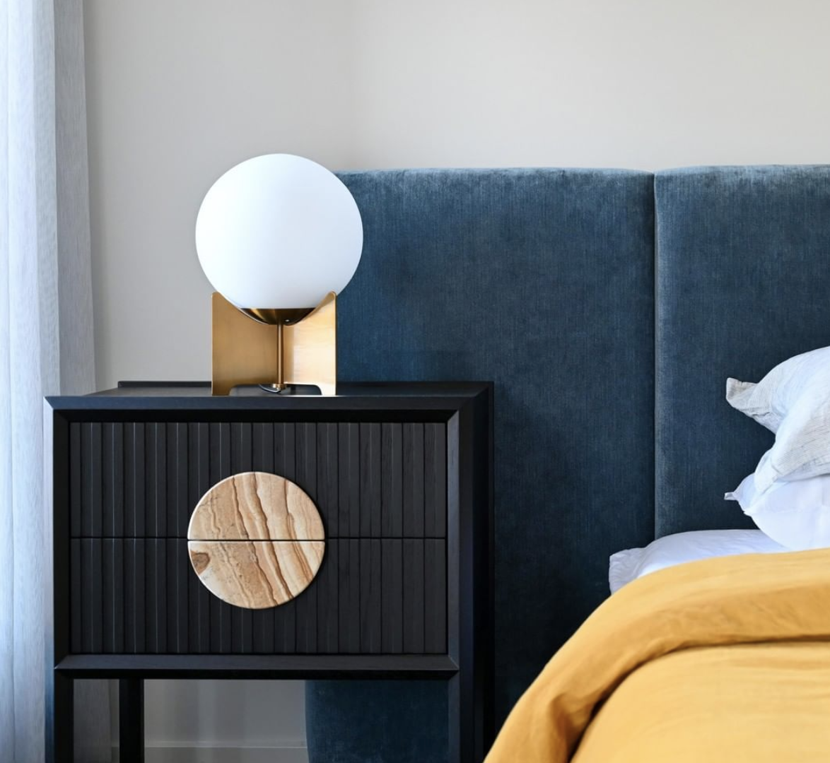 Globe table lamp on a black bedside cabinet beside a blue bedhead and mustard linen.