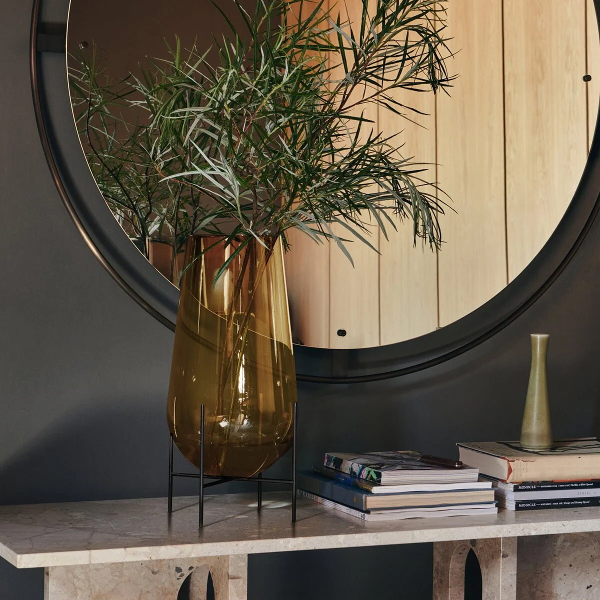 Amber glass vase with branches beside a round mirror and stacked books.