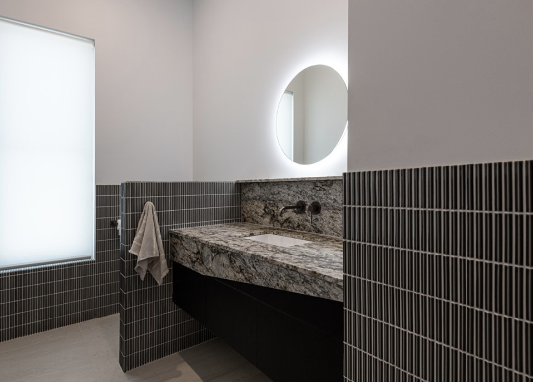 Stone bathroom vanity beneath a round illuminated mirror with dark wall tiles.