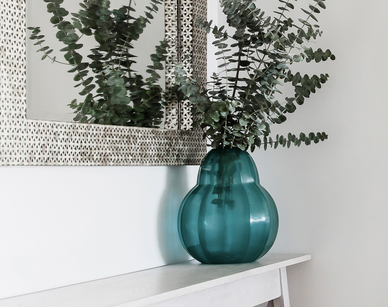 Teal glass vase holding leafy branches beneath a decorative mirror.