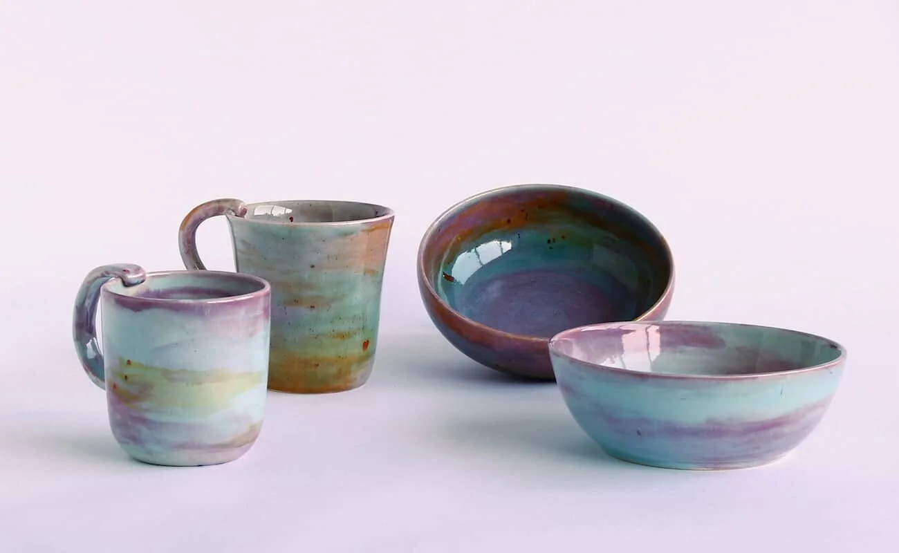 Colourful glazed ceramic cups and bowls displayed on a pink background.