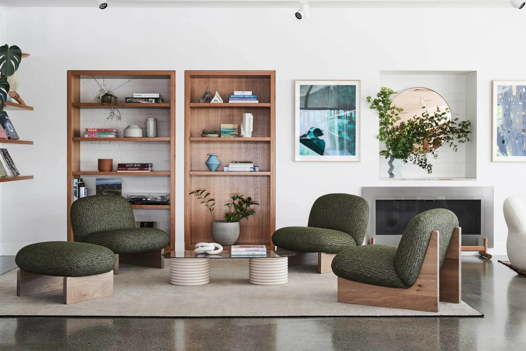 Green rounded armchairs and footstools beside timber shelves, framed art and flowers.