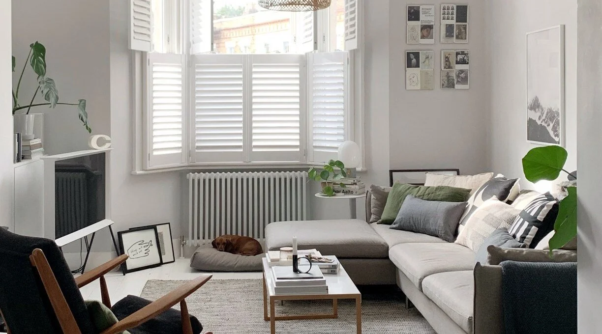 Living room with a pale sofa, black armchair, timber coffee table and window shutters.