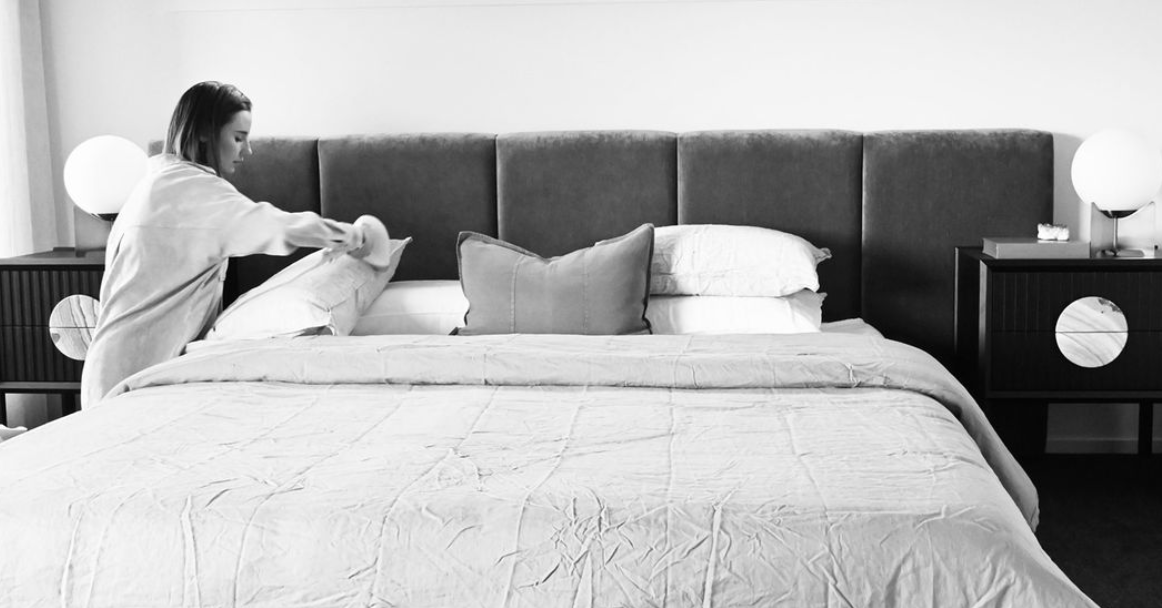 Black-and-white photograph of a person arranging pillows against an upholstered bedhead.