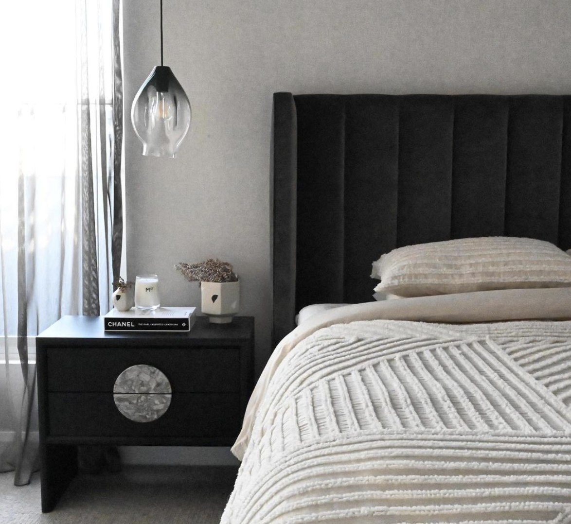Black channelled bedhead with pale striped linen, a dark bedside table and a glass pendant.