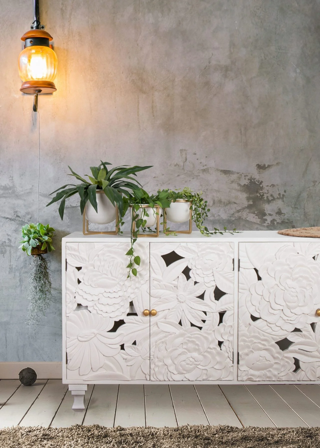 White patterned sideboard with plants and a warm wall light against a textured wall.