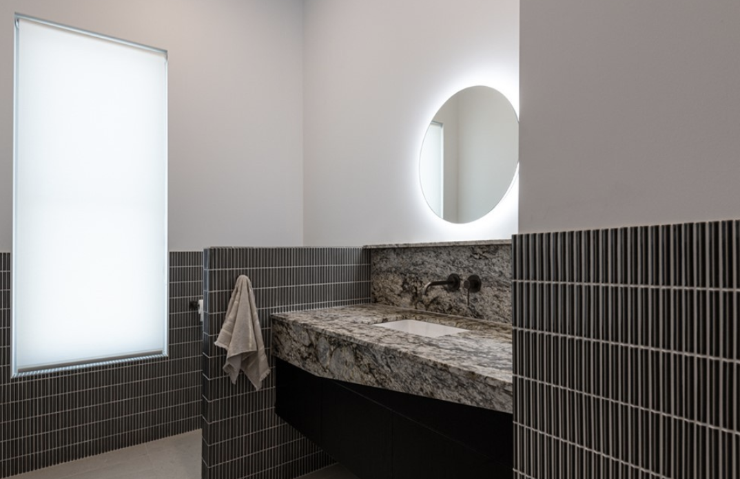 Stone vanity beneath a round illuminated mirror, with dark vertical tiles and a softly coloured wall.