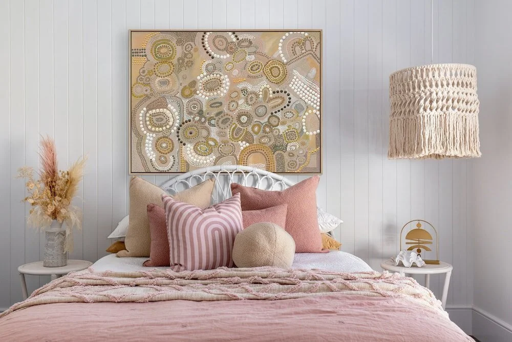 Bedroom with pink linen, decorative artwork, woven pendant lighting and layered cushions.