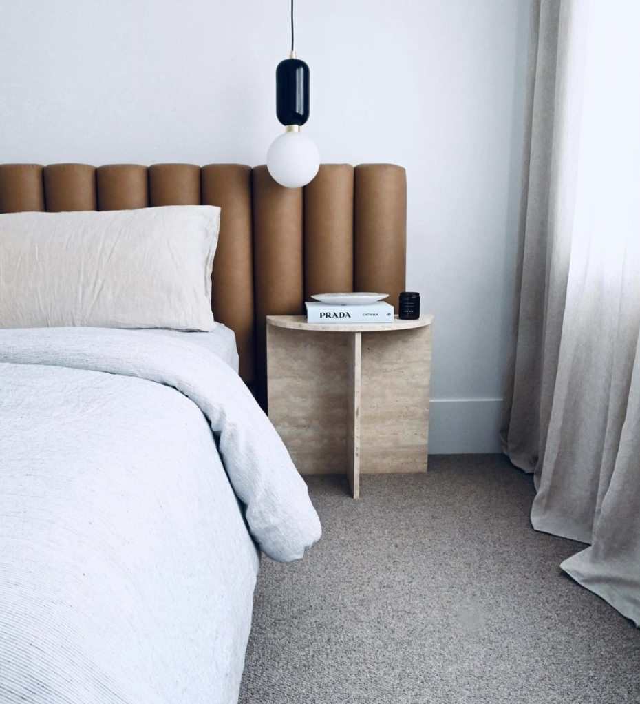 Tan channelled bedhead with white linen, a pale bedside table and a suspended light.