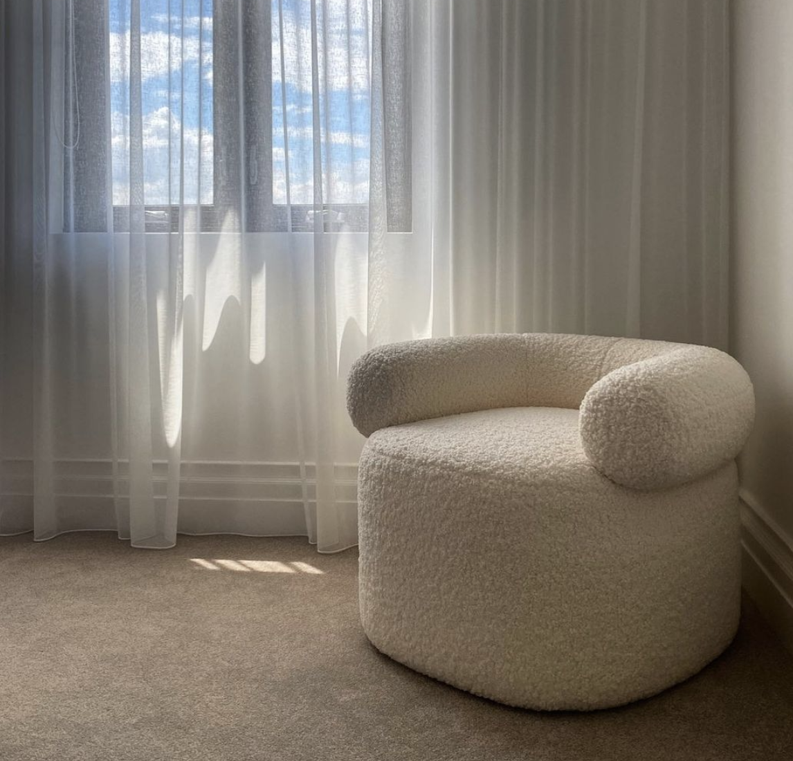 Rounded cream boucle armchair beside sheer curtains in a softly lit living room.