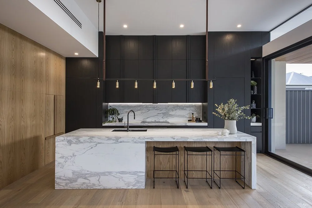 White marble kitchen island and splashback with grey veining, dark cabinetry, timber flooring and a linear pendant.