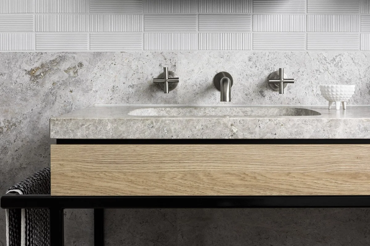 Pale limestone vanity with an integrated basin, timber drawer and wall-mounted cross-handle taps beneath textured tiles.