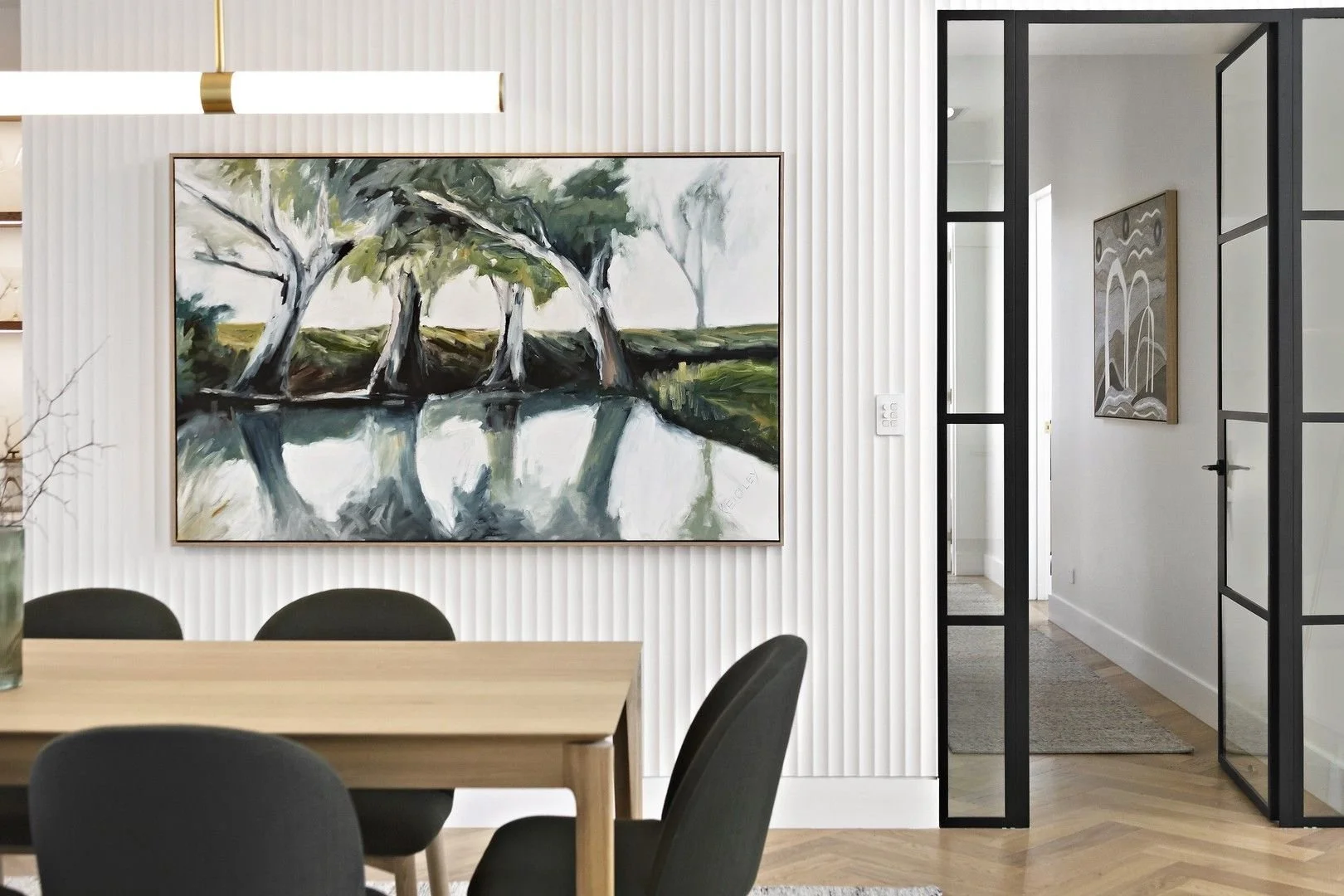 Dining area with framed landscape art, vertical wall panelling and black-framed glass doors.