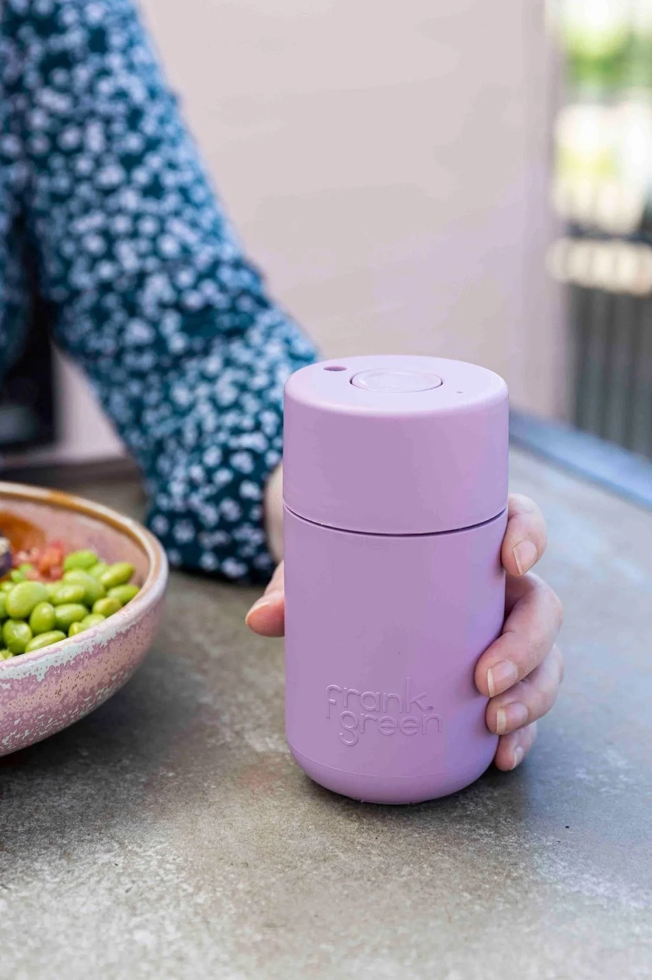 Lilac Frank Green reusable cup held above a grey tabletop.