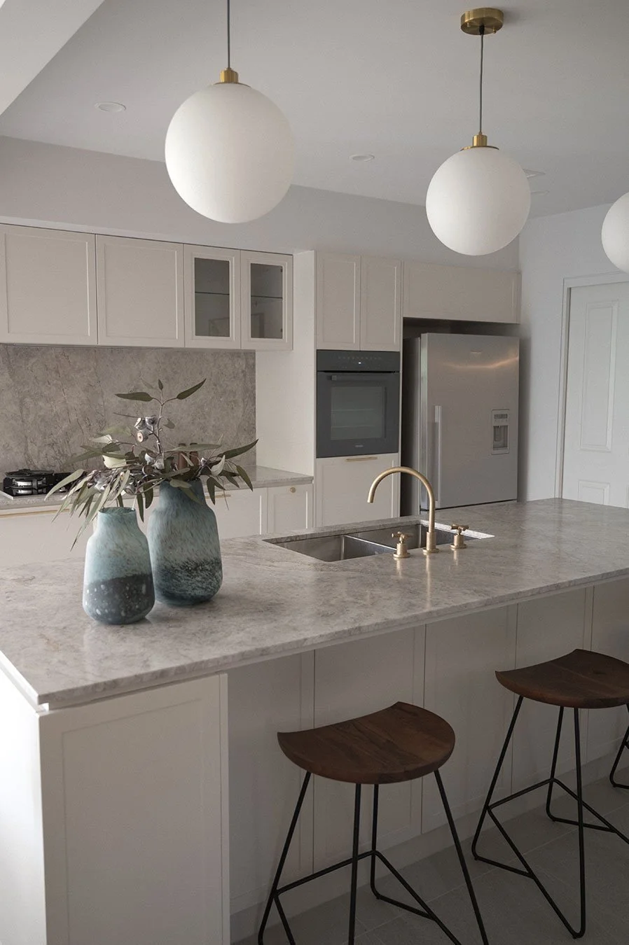 Modern kitchen with white cabinets, marble countertop island, two vases with greenery, gold faucet, and hanging globes lighting fixtures.
