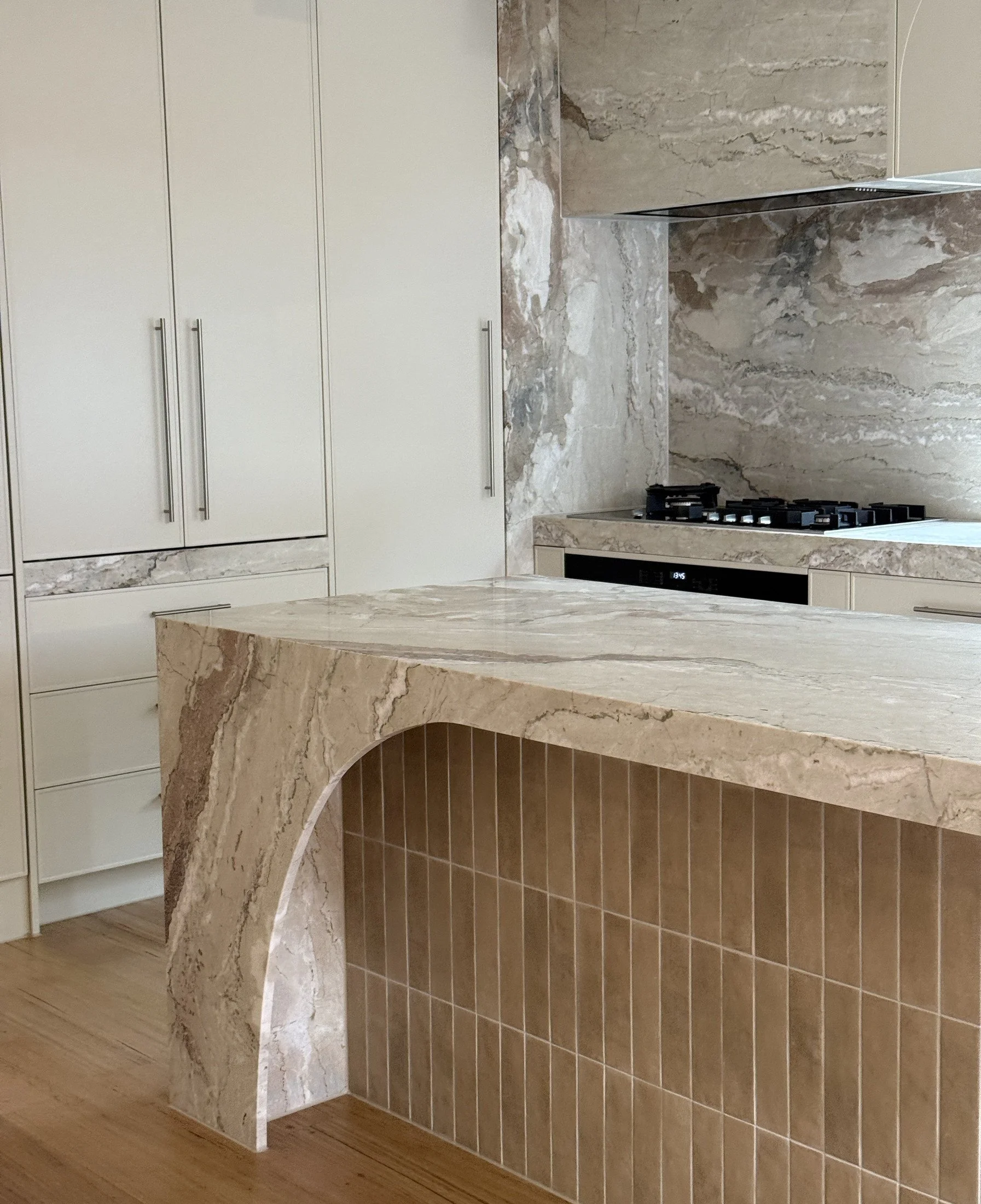 Kitchen with pale cabinetry, a veined stone island and arched detailing over vertical tiles.