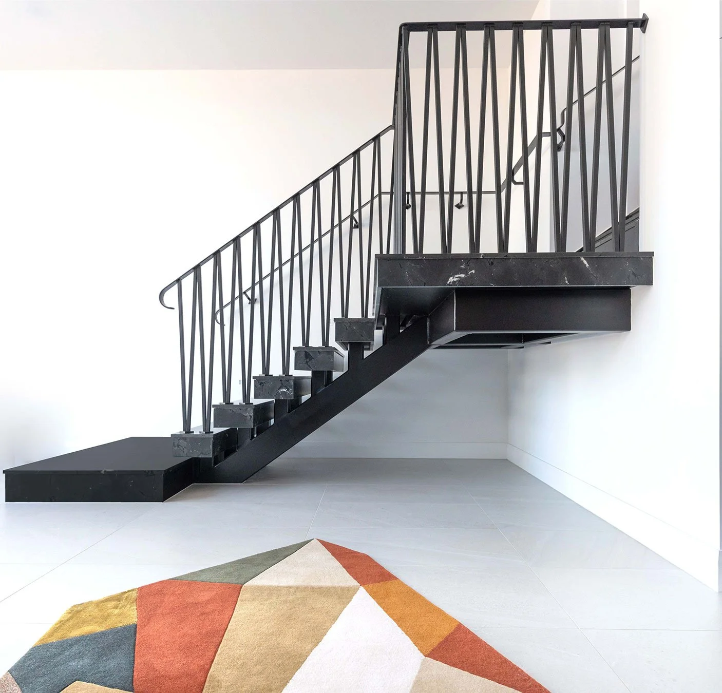 Modern black staircase with metal railings in a minimalist interior, adjacent to white walls and a colorful geometric rug on grey tile flooring.
