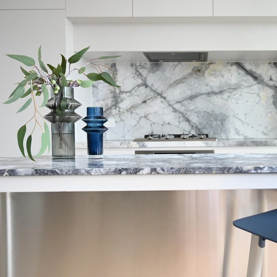 A kitchen countertop with a marble surface, decorated with two blue vases, one holding green leafy branches. The background features a marble backsplash and a stove. Part of a chair with a dark seat and light-colored legs is visible.