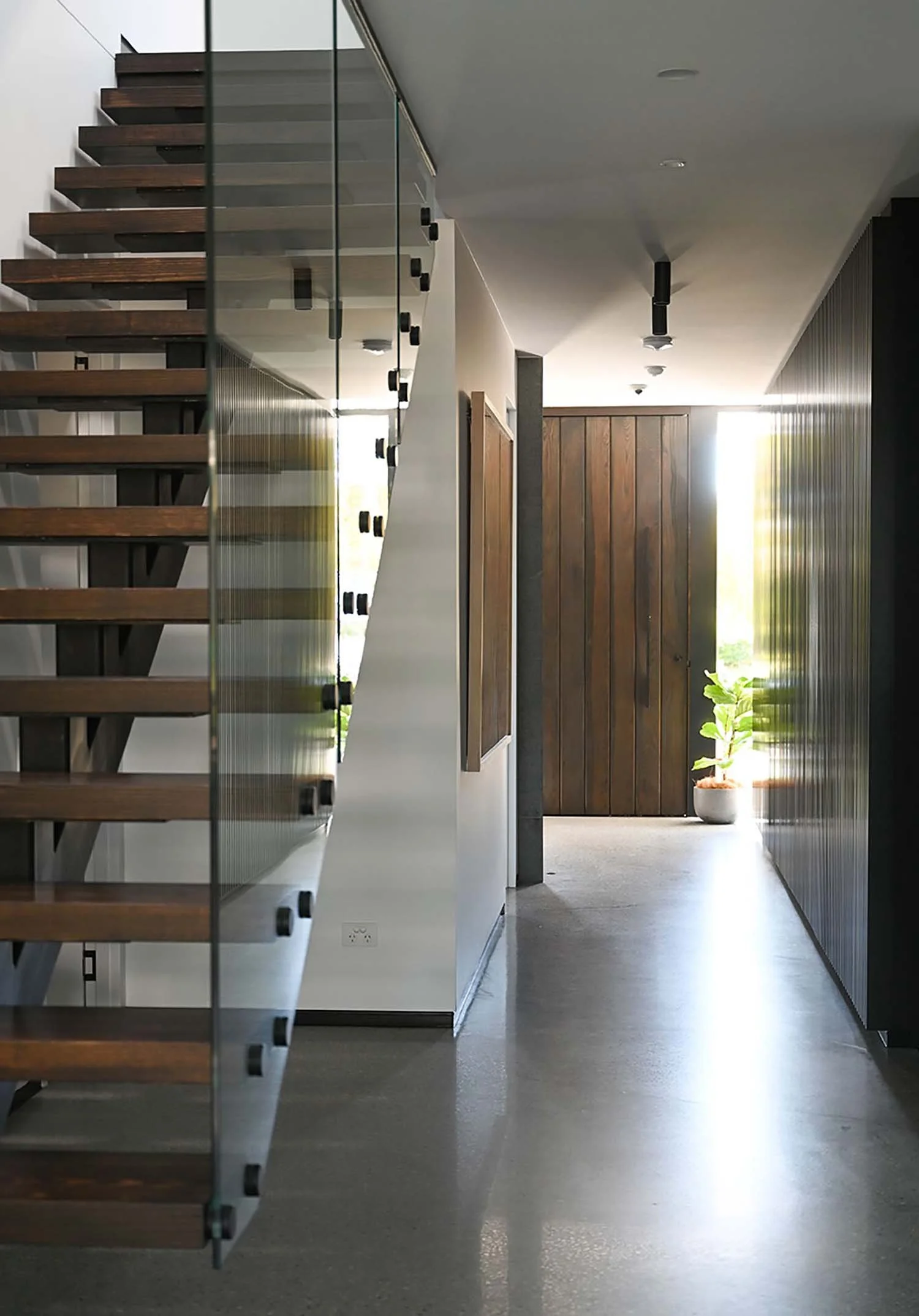 Interior hallway with floating wooden stairs on left, wooden wall on right, and a potted plant near the window at the end letting in natural light.
