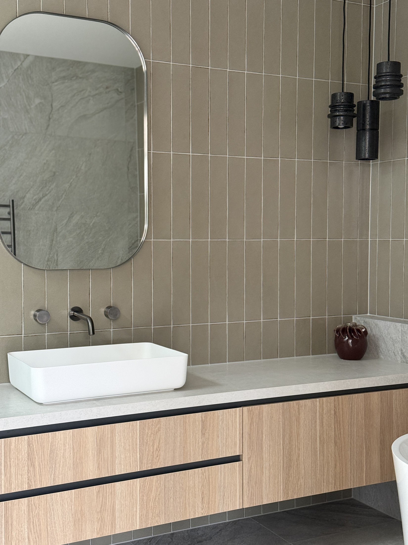 Timber bathroom vanity with a white vessel basin, rounded mirror, vertical tiles and black pendant lighting.