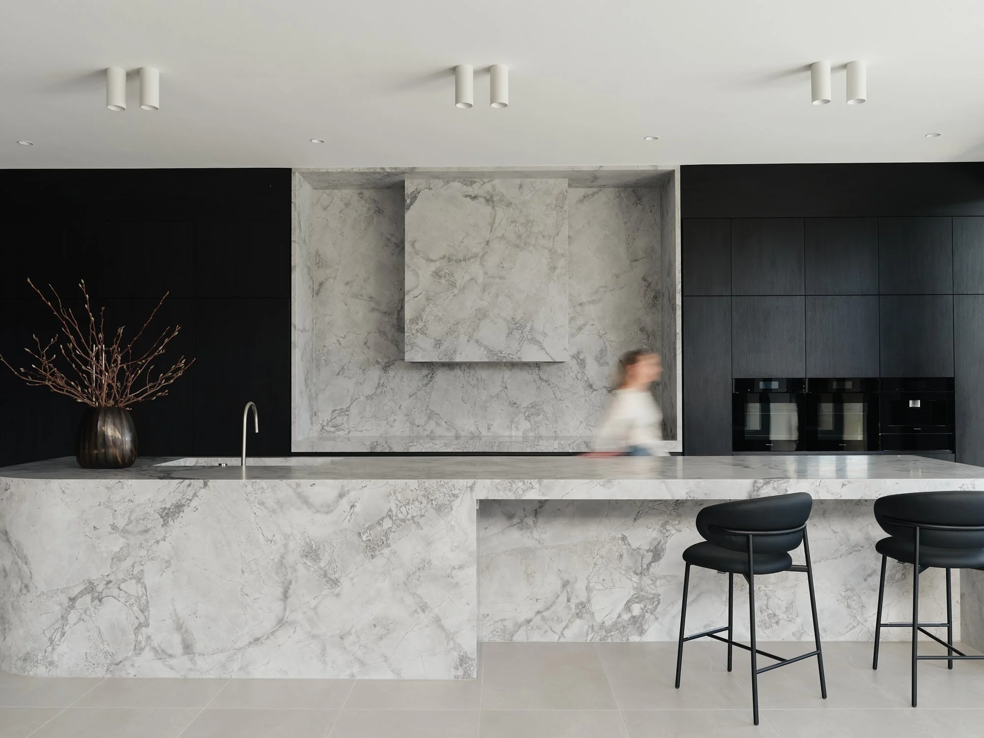 Wide view of the stone kitchen island, dark joinery and bar seating at Verde House.