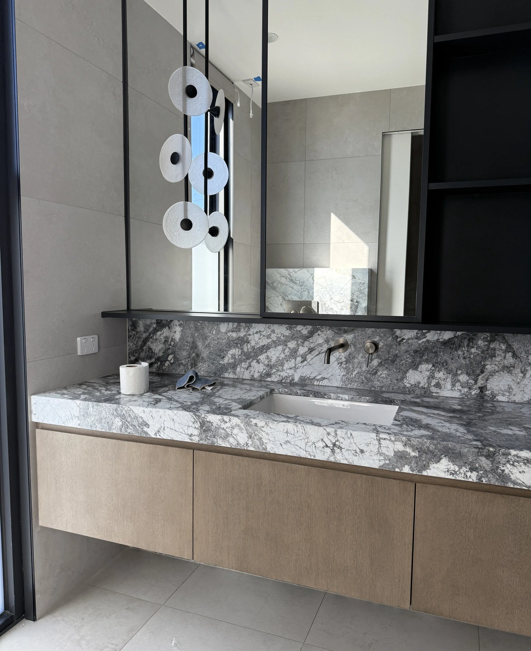 Timber bathroom vanity with a veined stone benchtop, large mirror and sculptural disc pendant light.