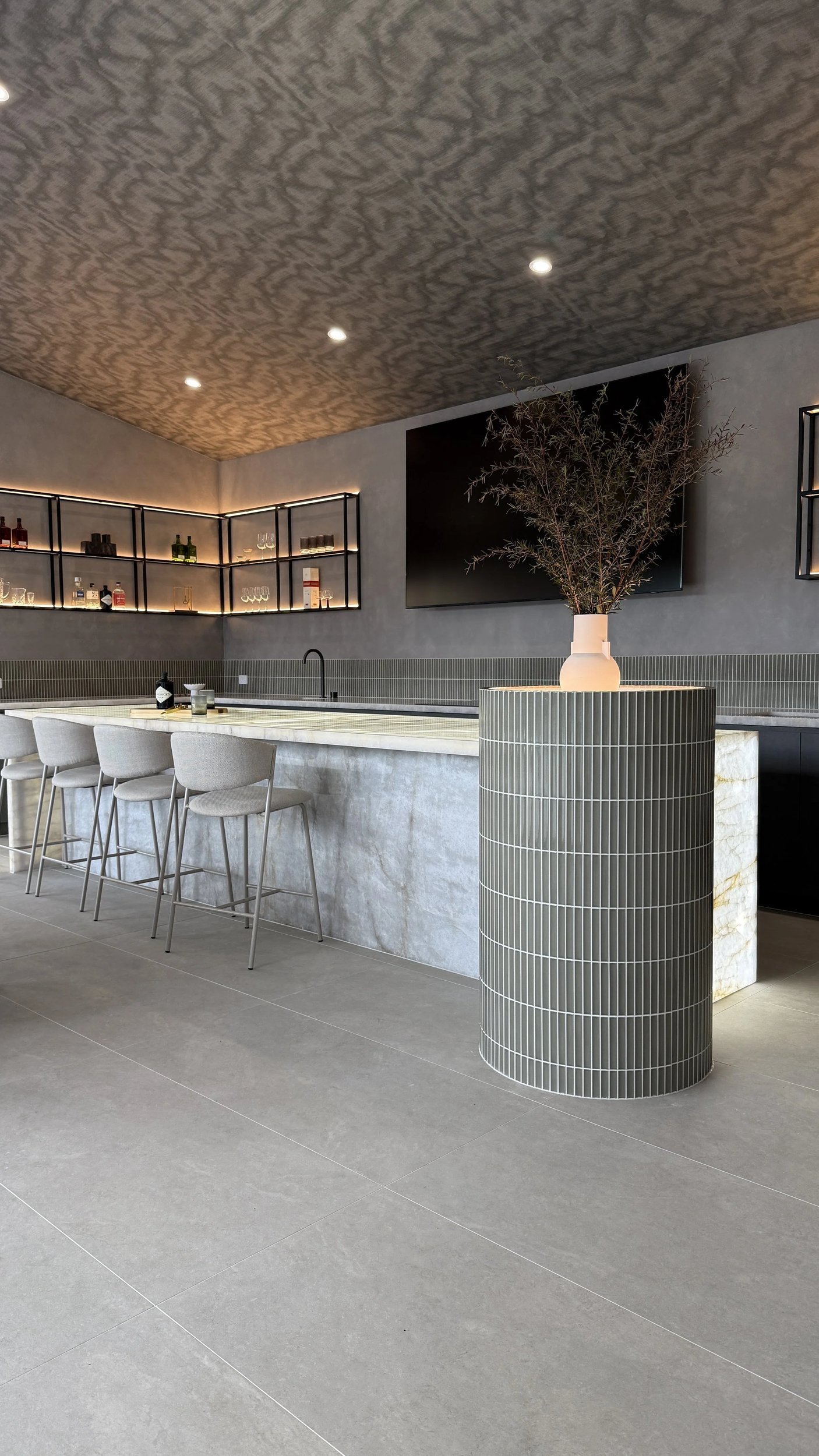 Home bar with a veined stone counter, high stools and dark cabinetry beneath illuminated shelves.
