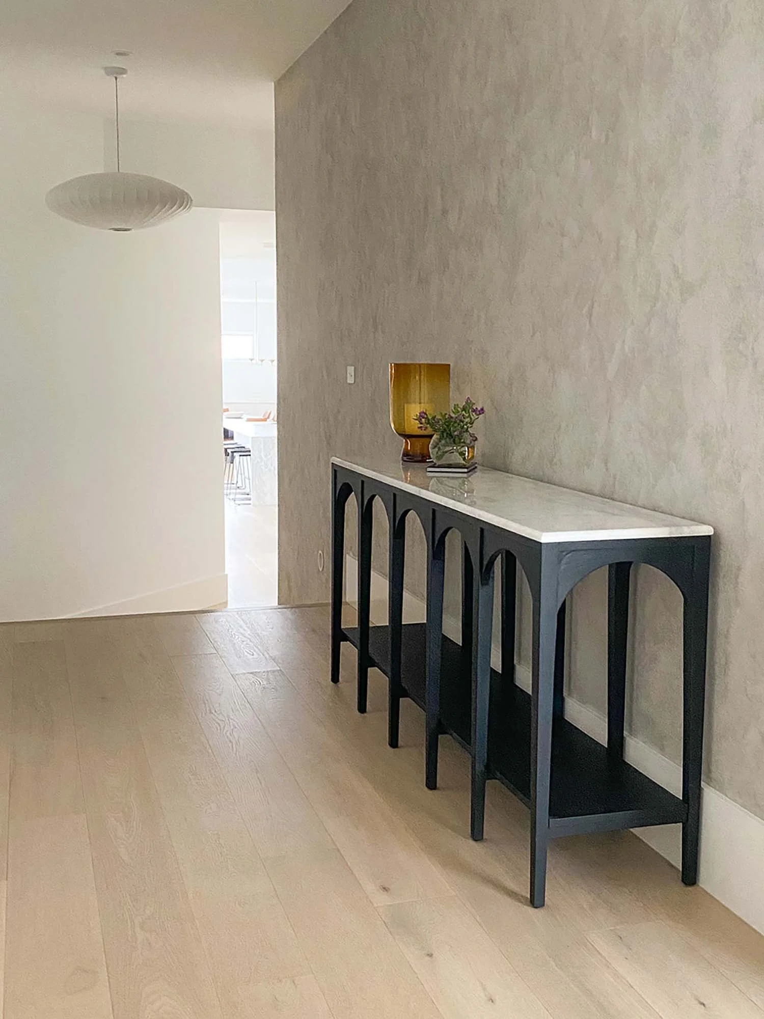 A black console table with a white marble top has a green glass vase with purple flowers and a yellowish decorative vessel on it. The table is against a textured beige wall in a room with light wooden flooring.