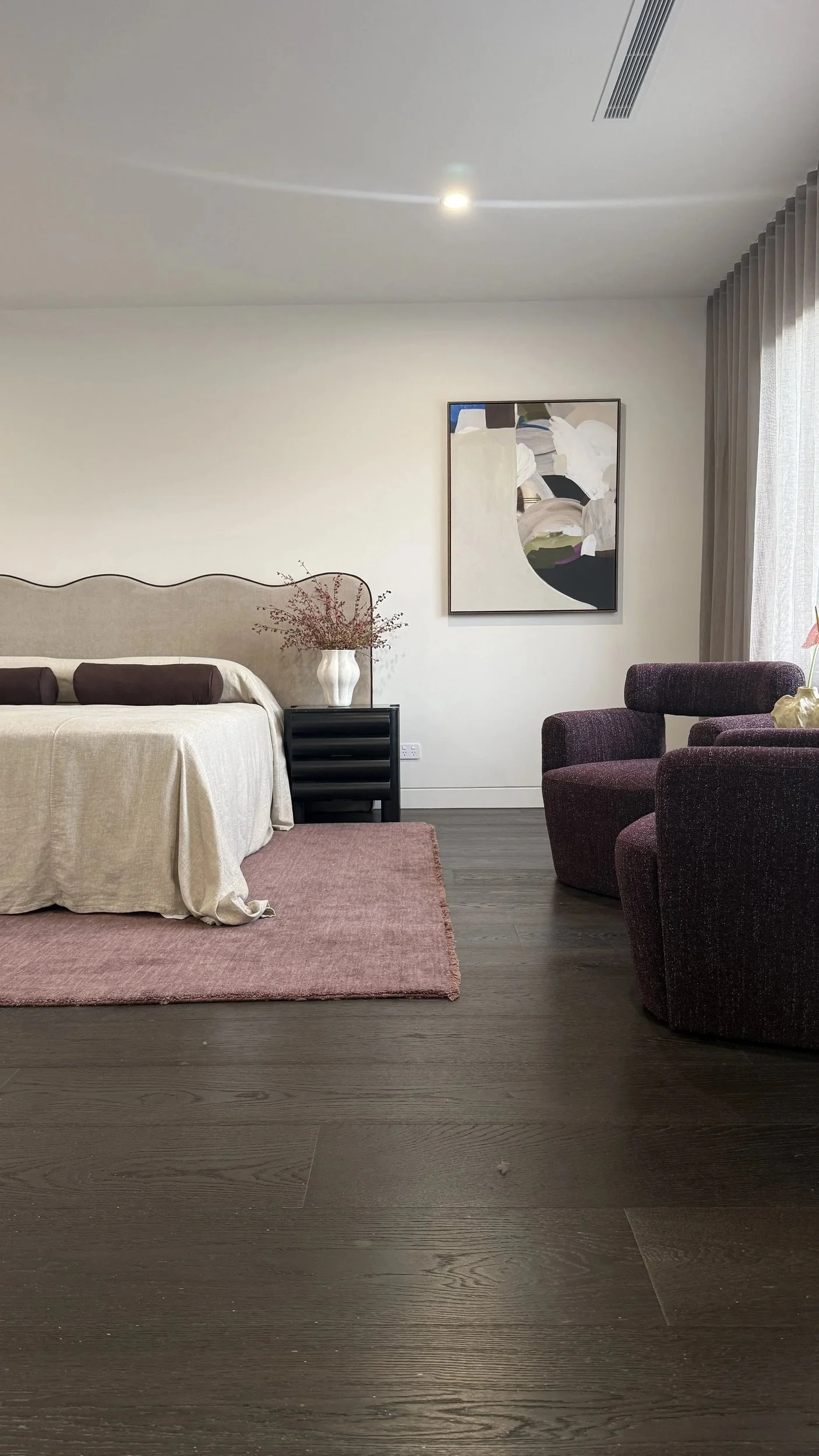 Bedroom with a curved upholstered headboard, pale bedding, a pink rug and burgundy chairs.