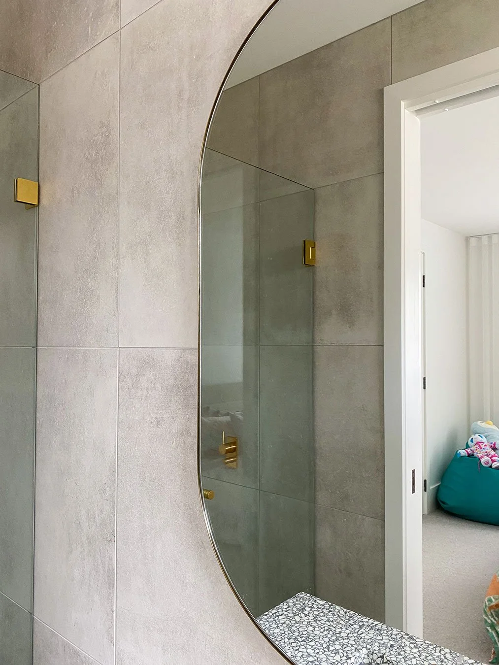 Close-up of a bathroom mirror reflecting beige tiled walls, gold fixtures, and a doorway leading to a room with a bean bag and stuffed animals.