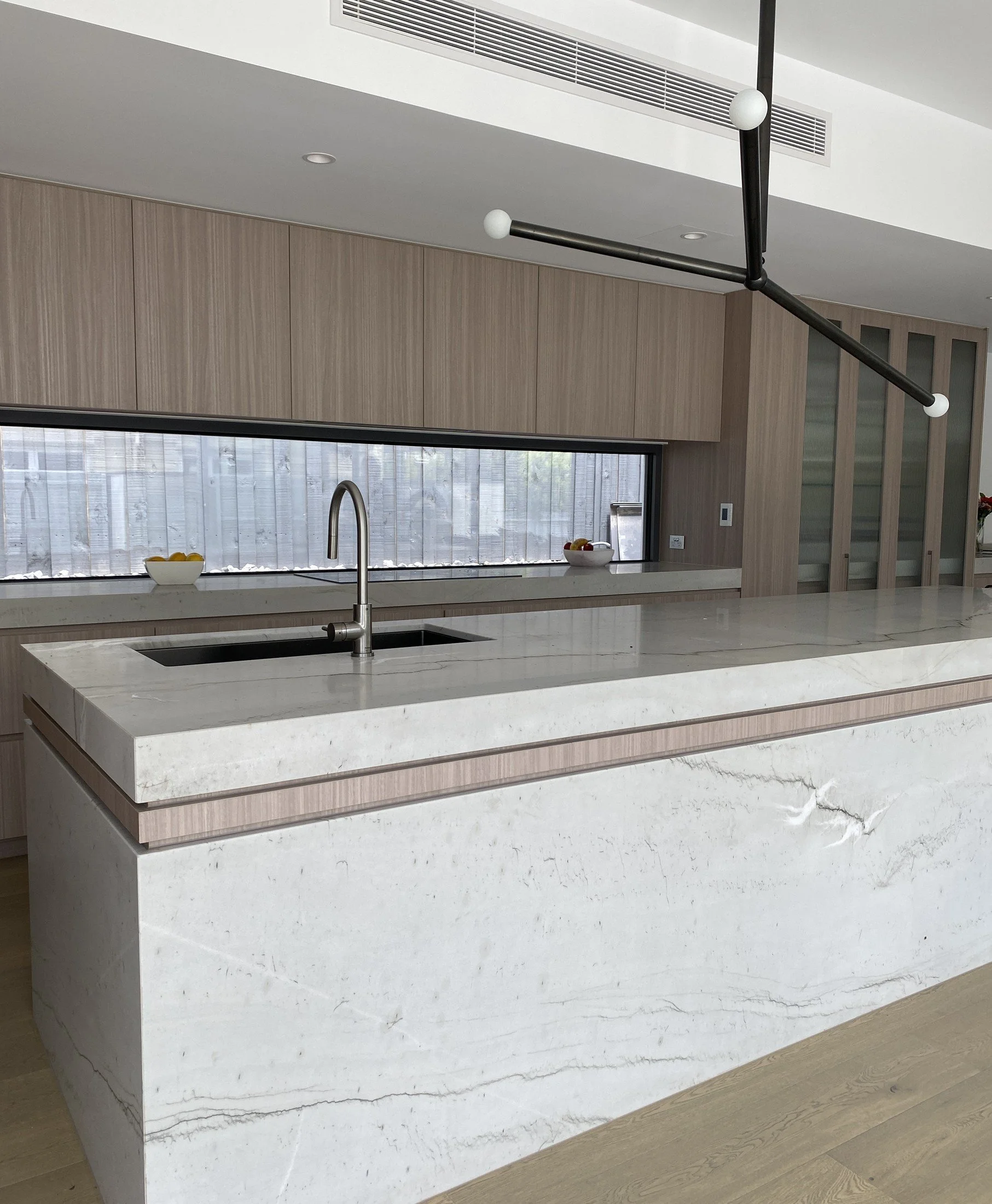 Timber kitchen with a pale veined stone island, integrated sink and suspended black linear light.
