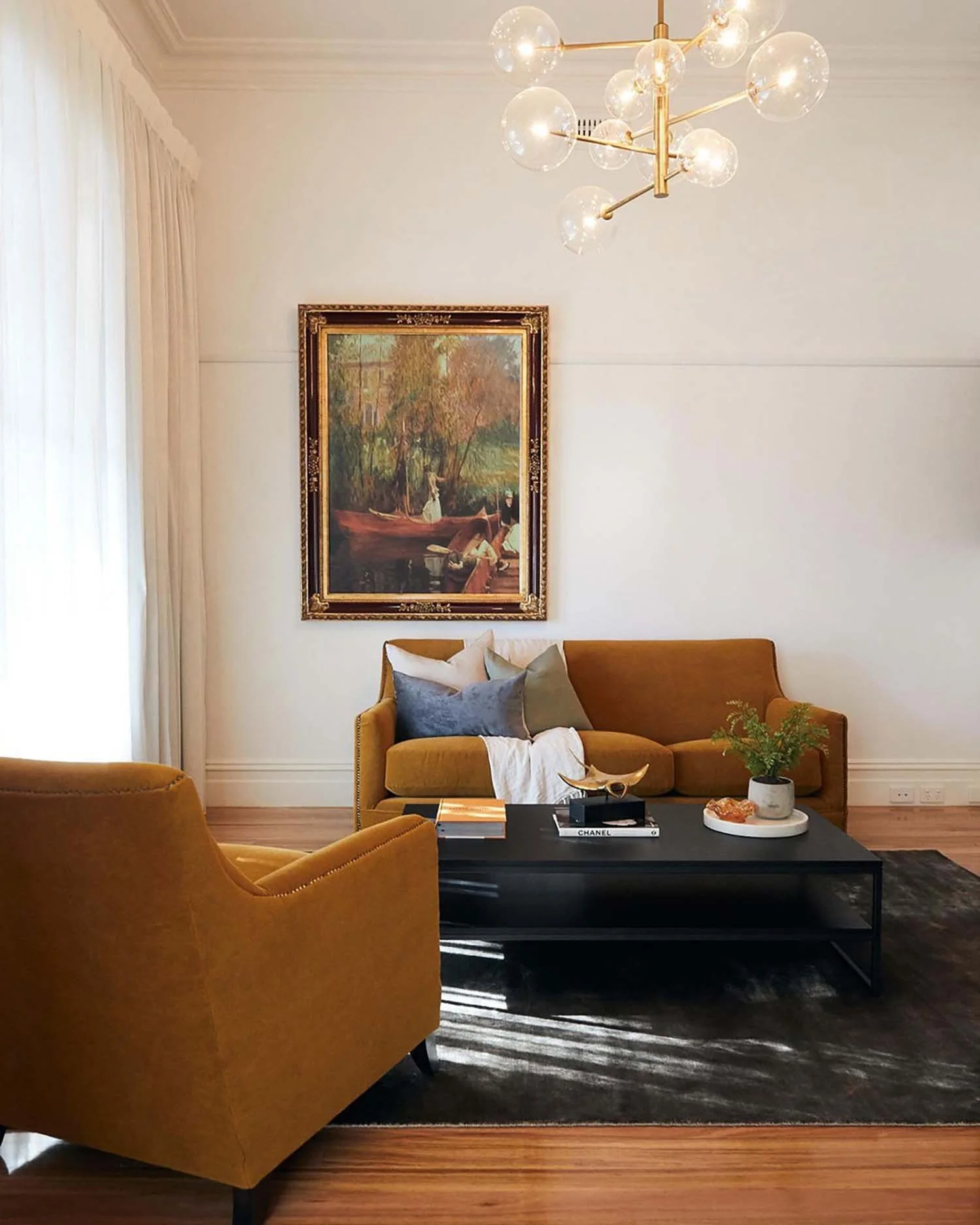A modern living room with mustard yellow armchairs, a black coffee table, a white couch with pillows, a painting on the wall, a chandelier with clear glass globes, a decorative plant, and books on the table.