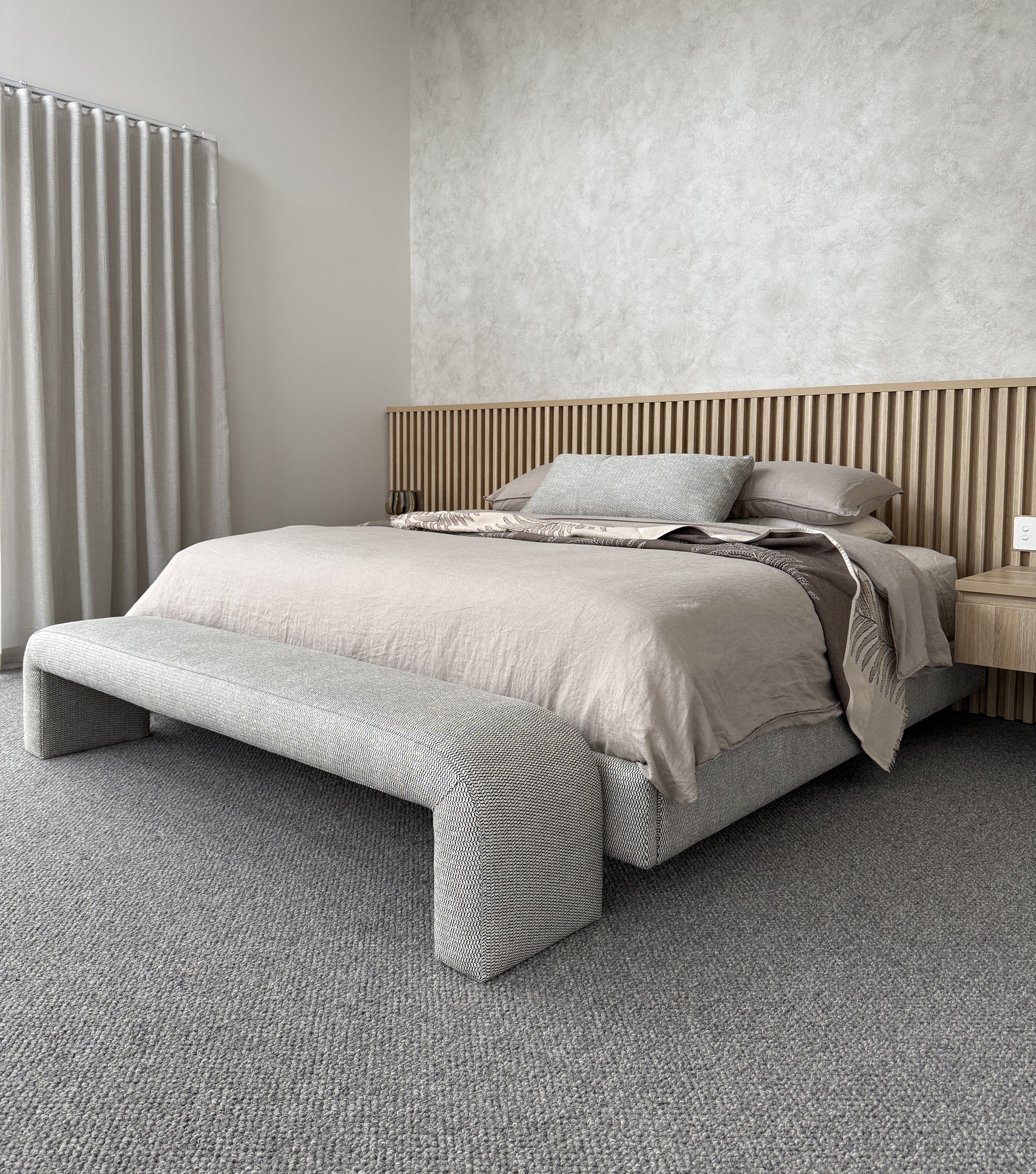 Bedroom with a slatted timber headboard, neutral bedding, upholstered bench and floor-length curtains.