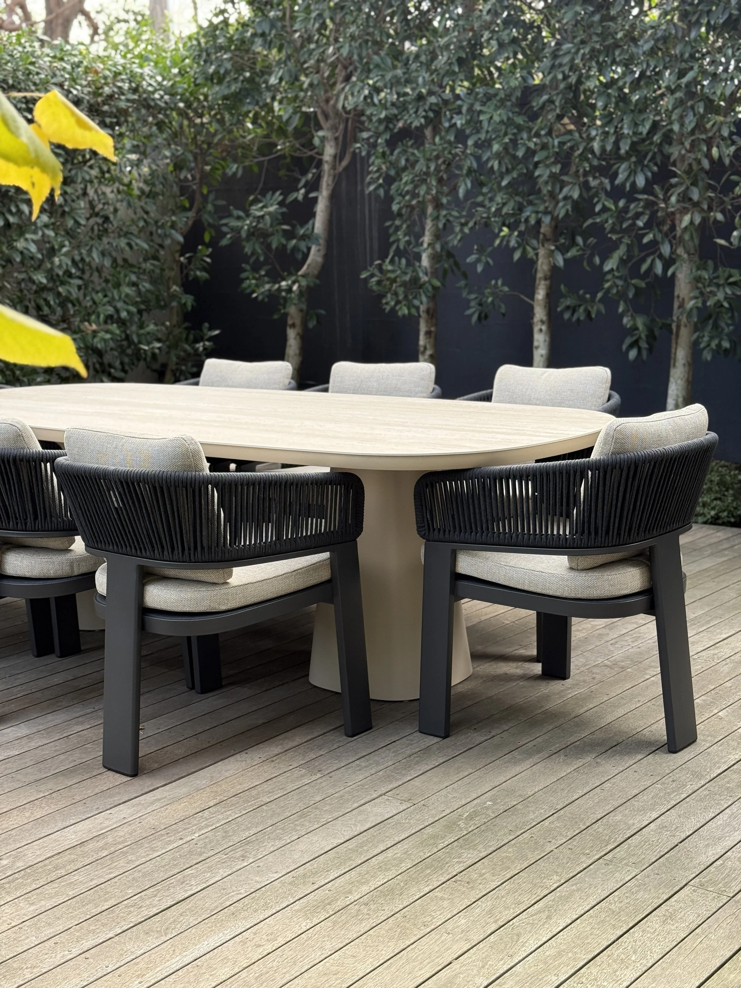 An outdoor patio table with black chairs and beige cushions, on a wooden deck, surrounded by green plants and trees.