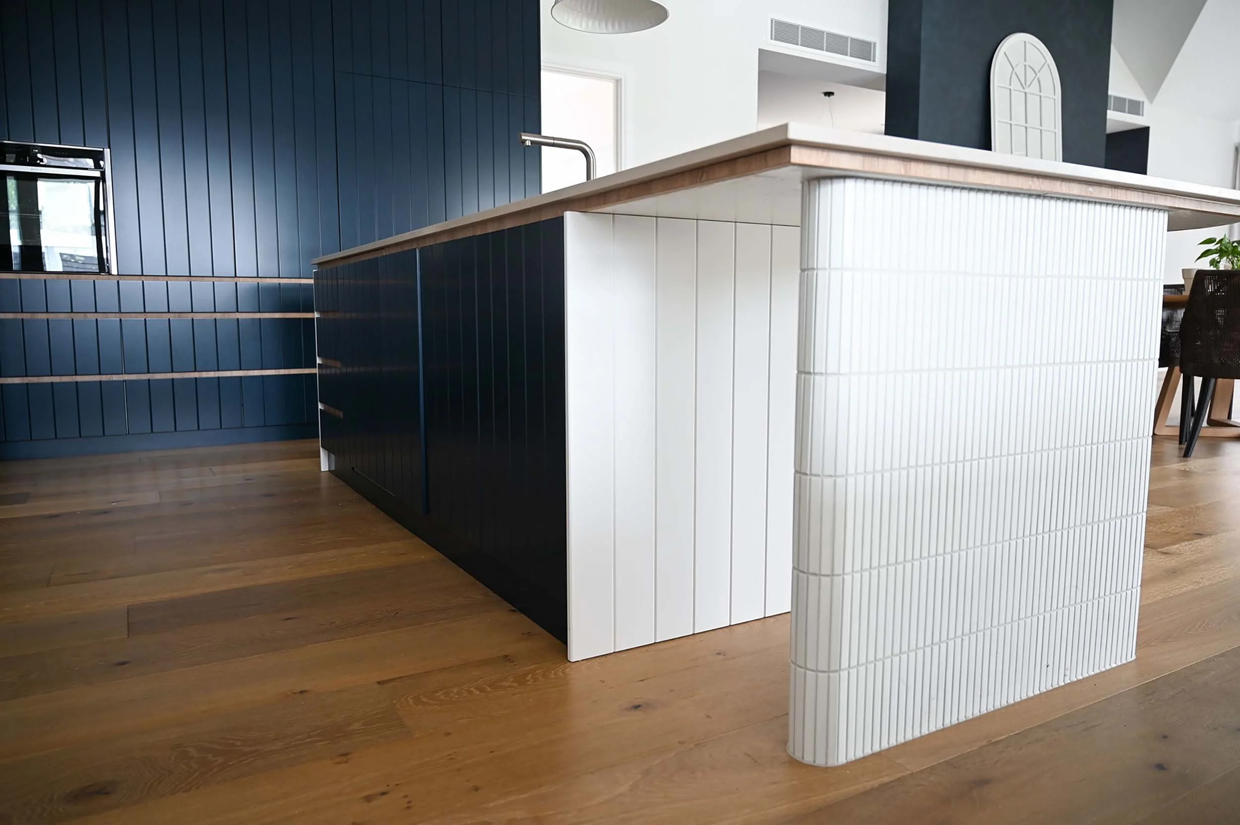 Modern kitchen island with white tile sides and a wooden top, situated on a wooden floor, with blue wall panels and a black chair nearby.