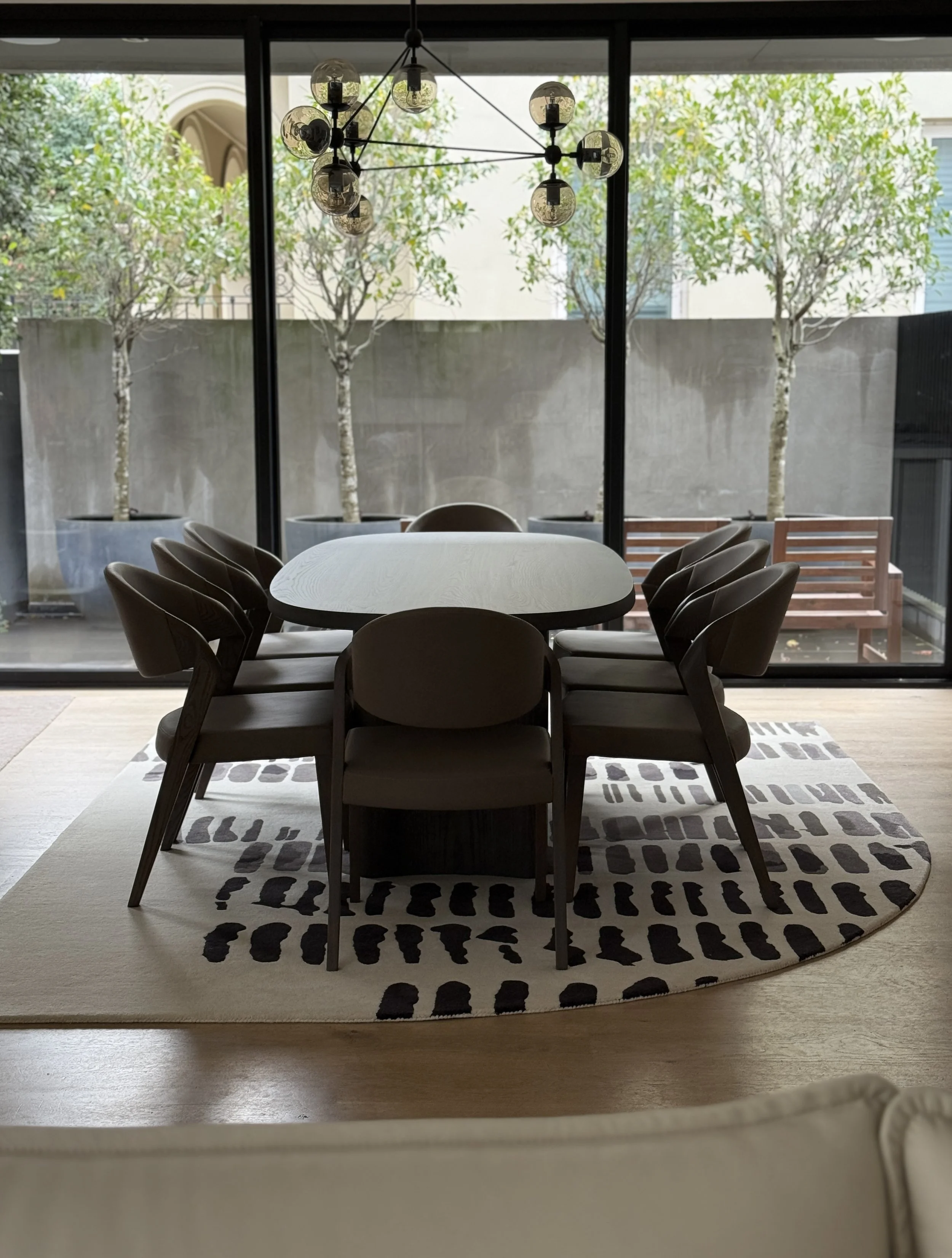 A dining area viewed from behind a beige upholstered chair, with a modern round table on a black and white spotted rug, through large glass sliding doors leading to an outdoor space with trees and a concrete wall.