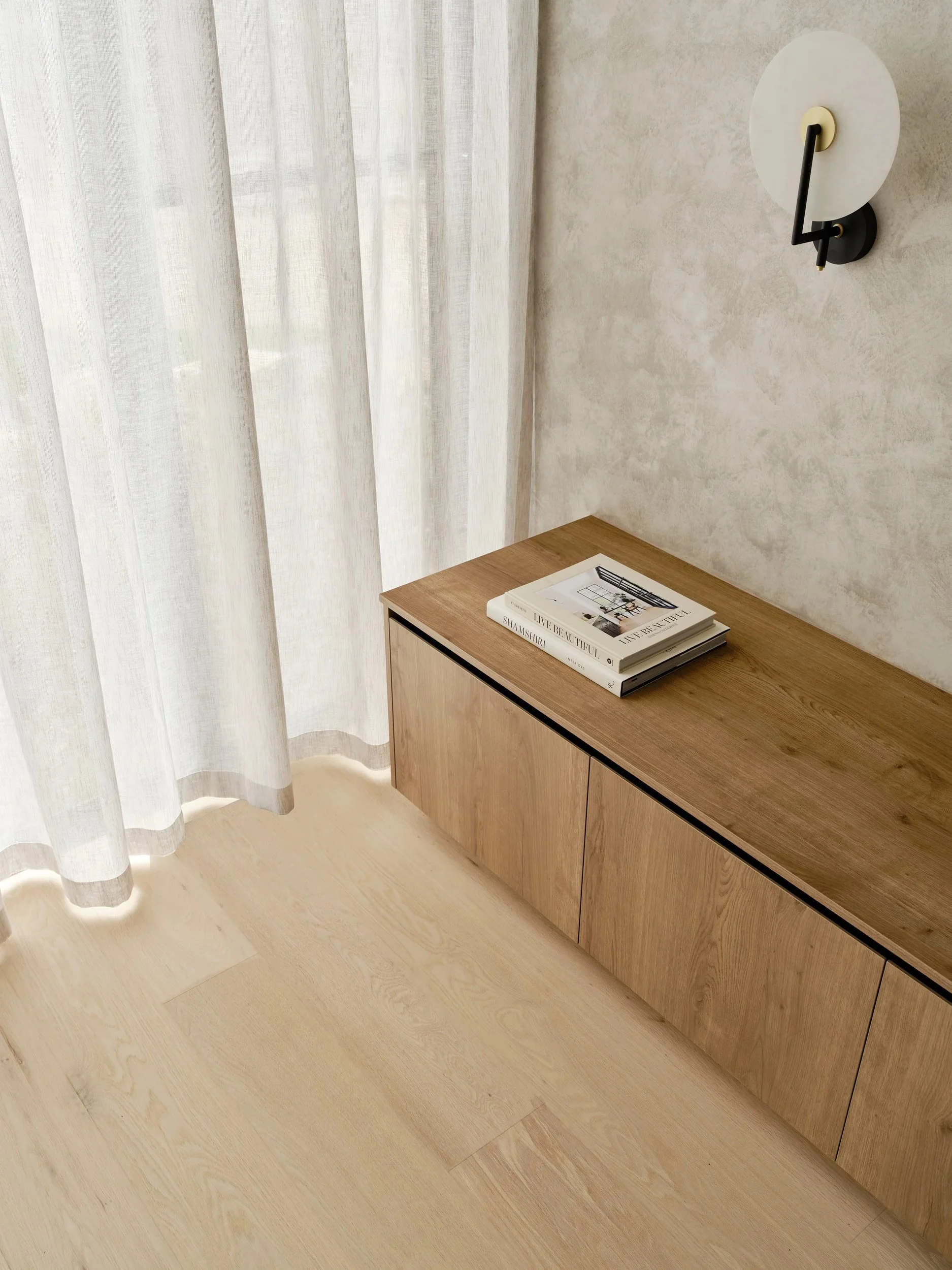Timber cabinet beneath a wall light beside sheer curtains and pale timber flooring.