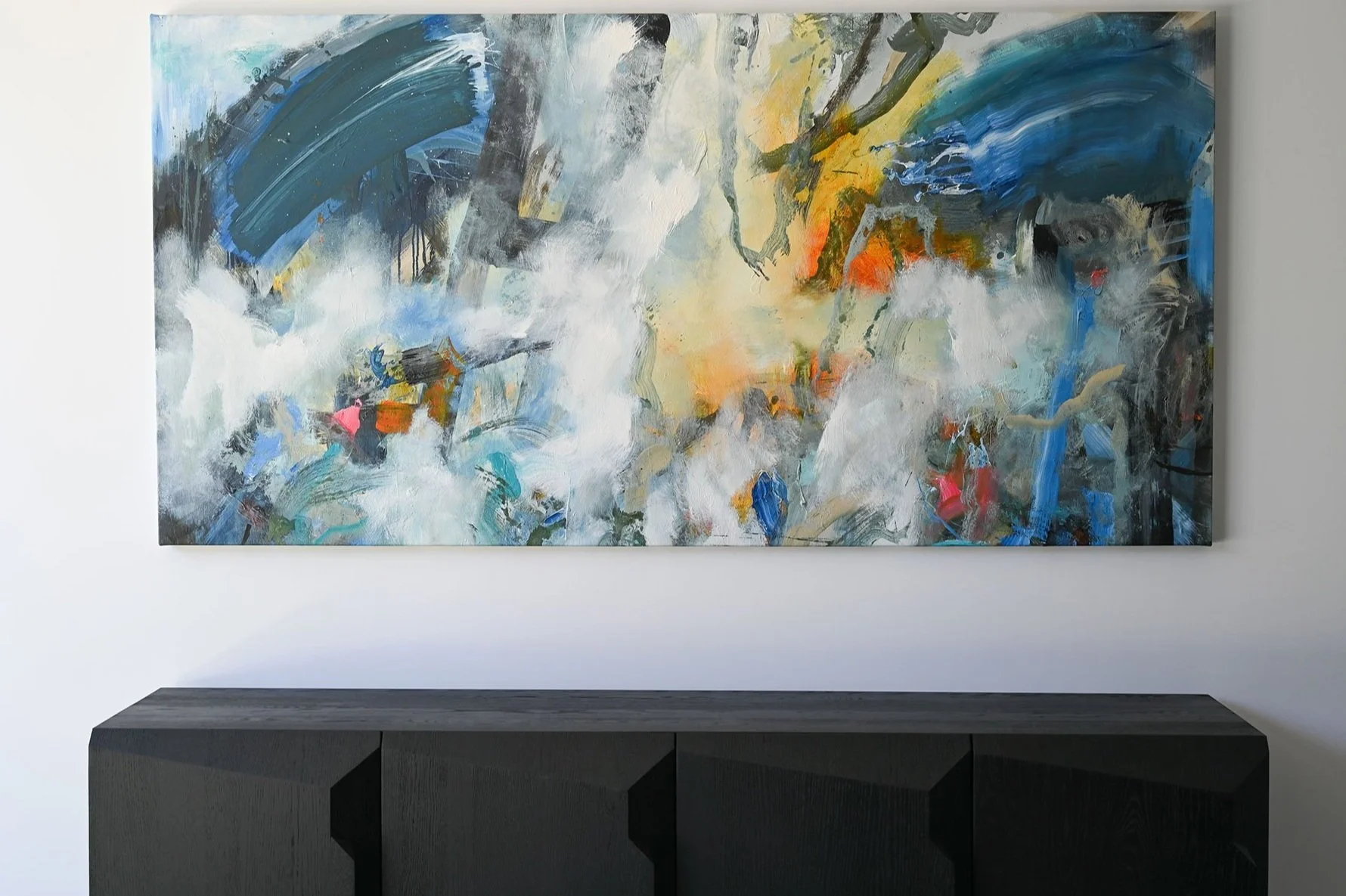 Abstract expressionist painting with bold brushstrokes of blue, white, yellow, orange, black, and pink, hanging on a white wall above a dark wooden console