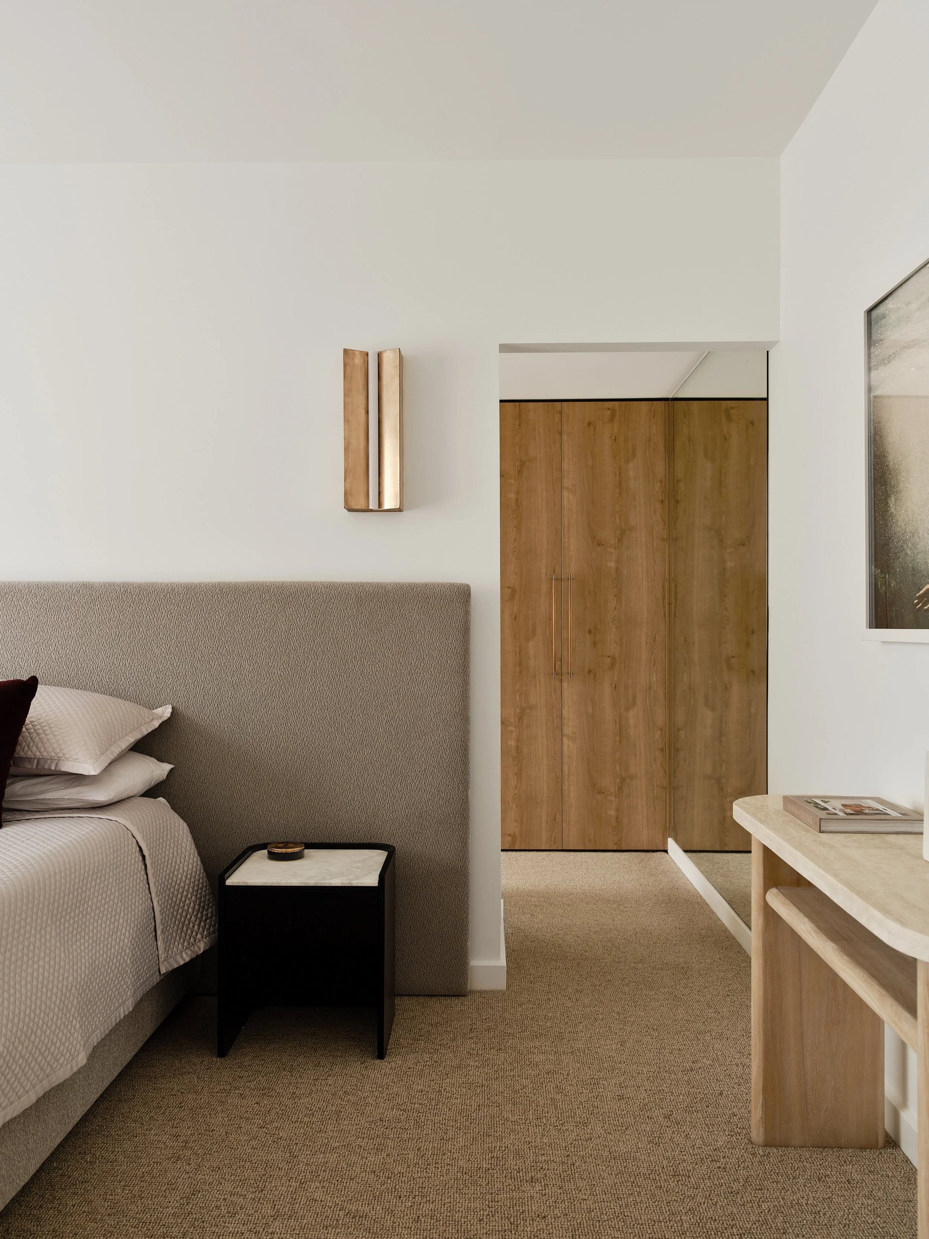 Bedroom detail with an upholstered headboard, dark bedside table and timber wardrobe beyond.