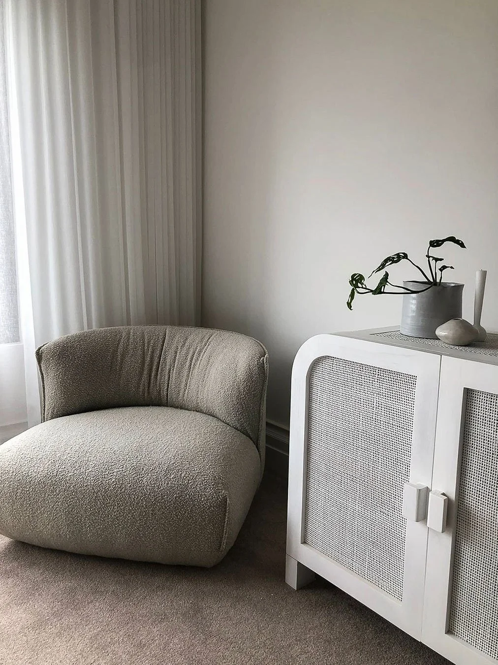 A cozy corner with a beige, textured armchair next to a white cabinet with rattan doors. On top of the cabinet, there are a dark gray vase with a leafless plant, a small white pot, and a tall white candle. Light filters in through sheer white curtain