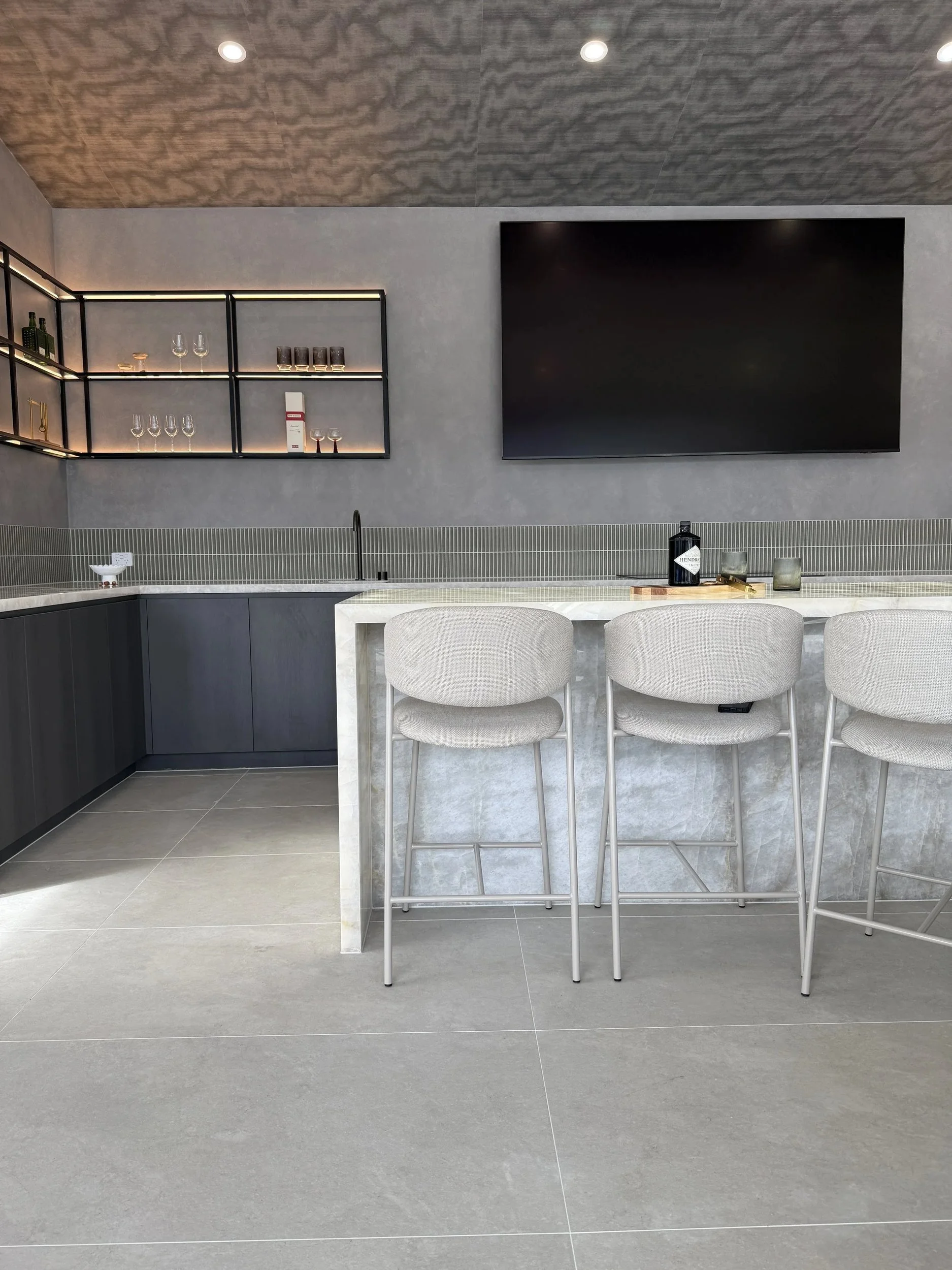 Wide view of the home bar with stone surfaces, upholstered stools and dark joinery.