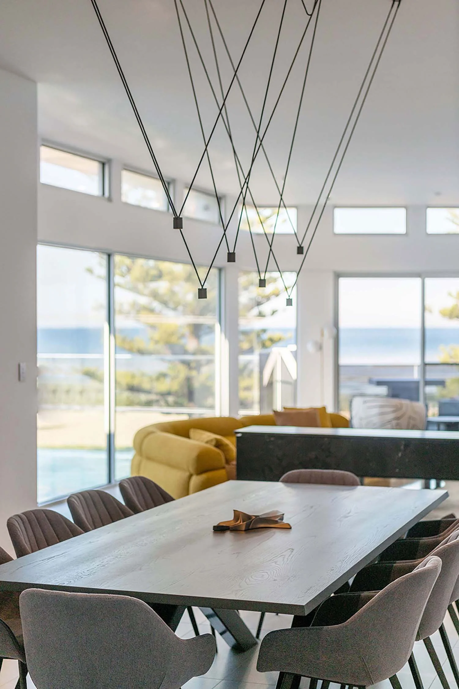 Modern dining room with a long wooden table surrounded by gray upholstered chairs, large windows bringing in natural light, a yellow lounge chair, and a contemporary ceiling light fixture with black cords and small square lights.