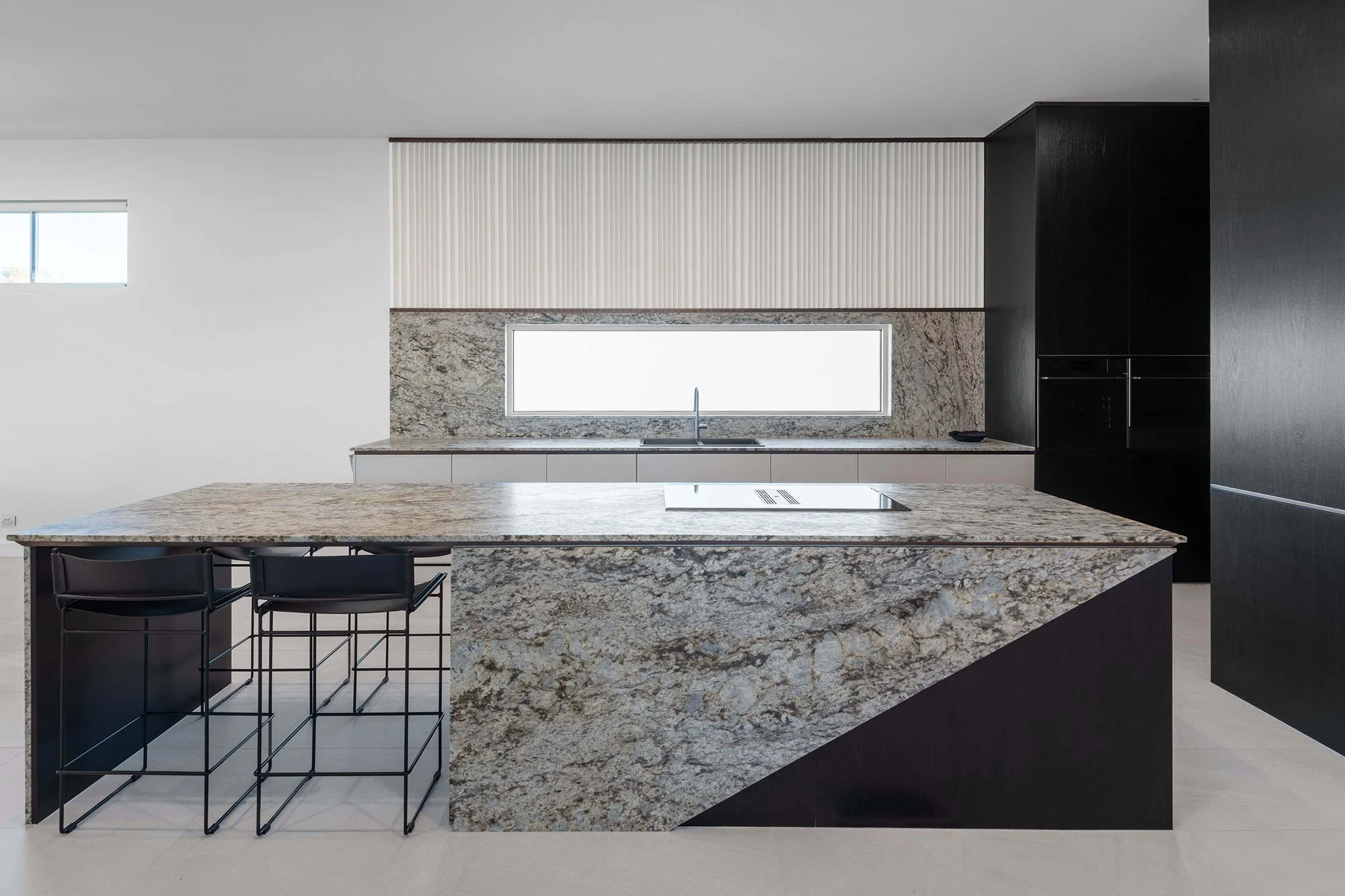 Modern kitchen with a large granite island, black chairs, a marble backsplash, a built-in sink under a high window, and high black cabinetry.