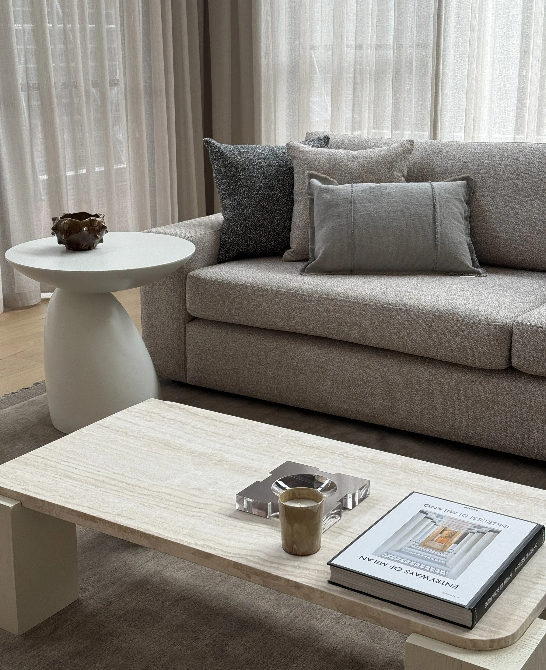 Neutral living room with a pale sofa, stone coffee table, sculptural white side table and full-height sheer curtains.