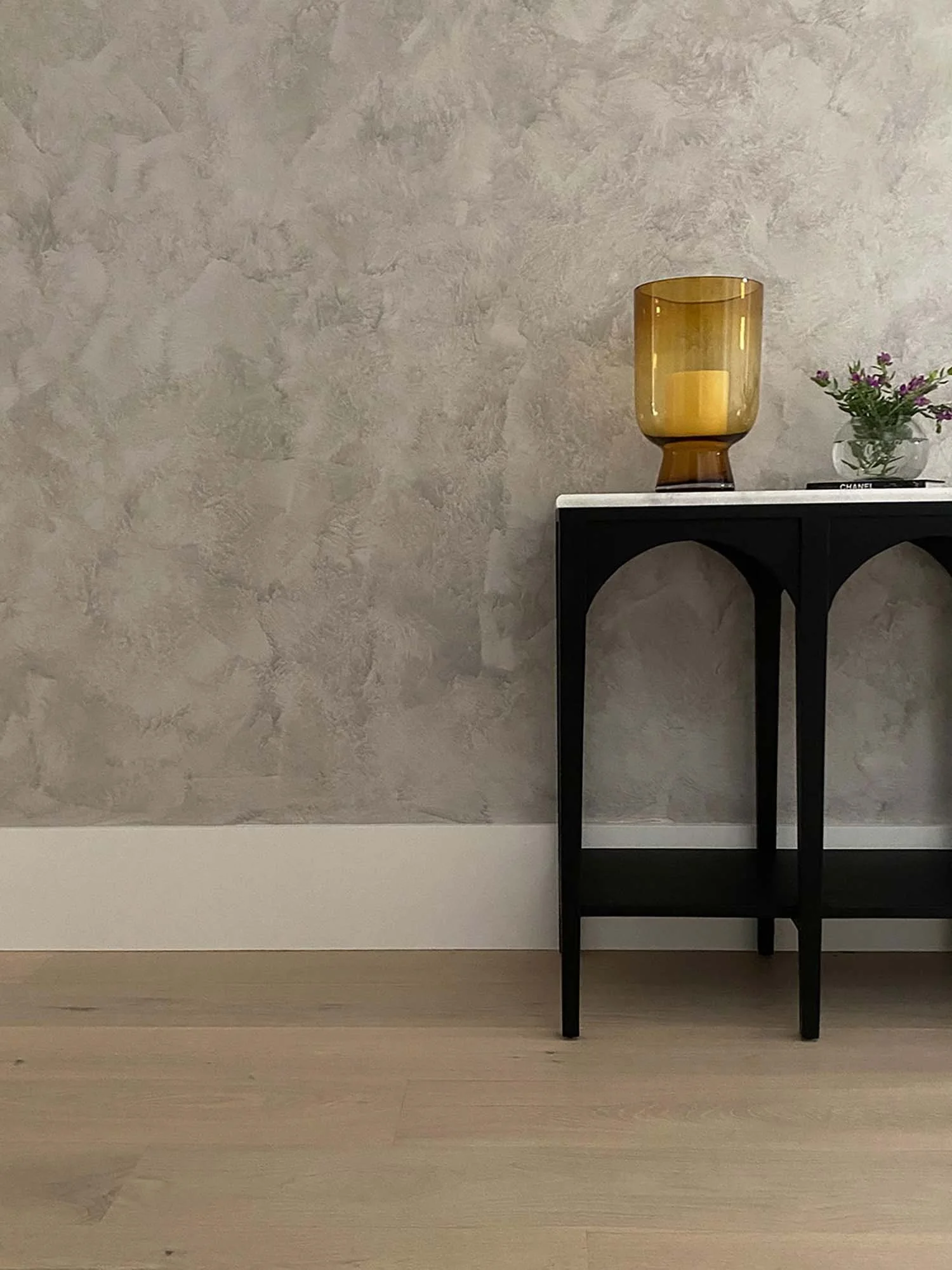 A black metal table with rounded edges on a light wooden floor and a textured beige wall in the background. On the table, there is a large amber-colored glass vase, a small clear glass bowl with purple flowers, and a book titled "CHANEL."